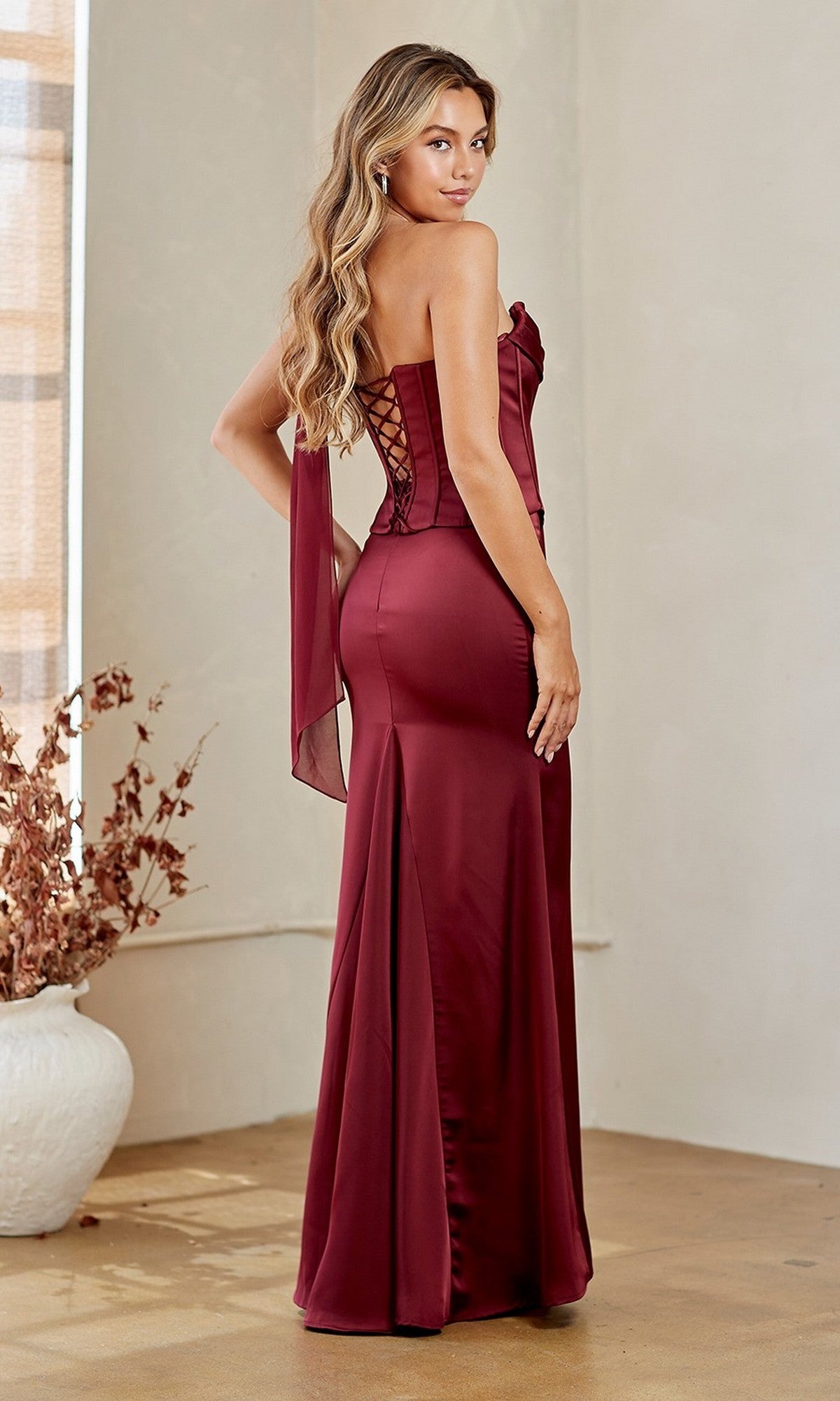 A woman in the Amelia Couture 6138 long prom dress, a strapless burgundy gown with a corset bodice and lace-up back, stands indoors on a polished floor beside a white vase with dried foliage.